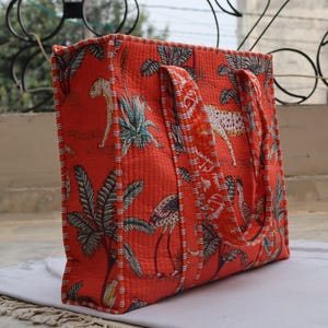 May include: Quilted tote bag in a vibrant orange colour, featuring a repeating pattern of leopards, tropical plants, and birds. The bag has sturdy handles and a quilted texture, offering a stylish and functional design for carrying essentials.