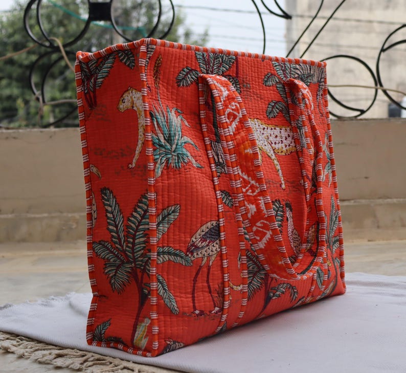 May include: Quilted tote bag in a vibrant orange colour, featuring a repeating pattern of leopards, tropical plants, and birds. The bag has sturdy handles and a quilted texture, offering a stylish and functional design for carrying essentials.