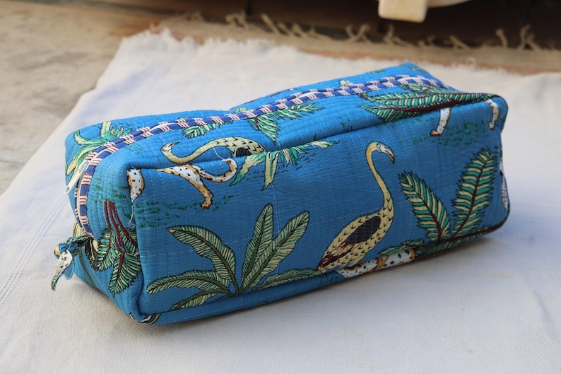 May include: A rectangular, blue fabric pouch with a tropical bird and foliage pattern. The pouch features a zipper closure and is likely used for storing cosmetics or small items. The design includes a mix of green, yellow, and white accents.