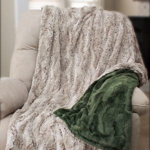 May include: A plush, reversible blanket draped over a beige armchair. One side is a light brown, textured faux fur, and the other is a deep green, smooth fabric. The blanket's soft texture and color palette create a cozy, inviting feel.
