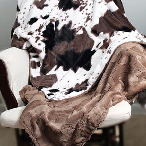 May include: A soft, plush throw blanket with a brown and white cowhide print on one side and a solid brown faux fur on the other side. The blanket is draped over a chair.