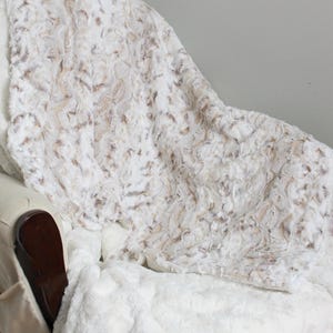May include: A plush white and tan faux fur blanket with a swirling, textured pattern draped over a chair. The top right corner features a small logo with two stylized elephants and the text "kemaily" in a simple, dark font.