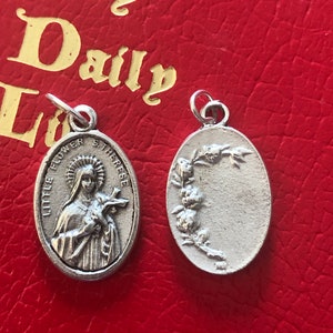 May include: Two silver oval-shaped medallions. One features a depiction of Saint Therese of Lisieux, with the text "Little Flower" and "St. Therese". The other medallion has a floral design.