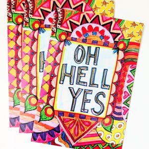 May include: A stack of colorful art prints with the phrase "OH HELL YES" in bold letters. The prints feature vibrant patterns in red, pink, yellow, and green, with geometric and floral designs. The overall theme is artistic and expressive.