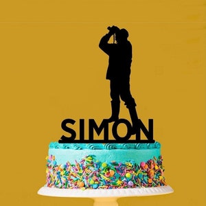 May include: A cake with blue icing and colourful sprinkles, topped with a black silhouette of a person holding binoculars and the name "SIMON". The cake is on a white stand against a yellow backdrop.