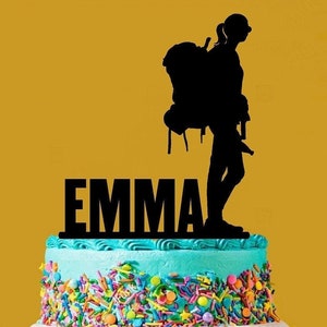 May include: A black silhouette of a person with a backpack on top of a blue and white cake with colourful sprinkles. The name "EMMA" is written in black letters on the cake.