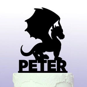 Op de afbeelding: Zwarte silhouet van een draak met uitgespreide vleugels, staand op een witte taart met de naam "PETER" eronder in zwarte letters.