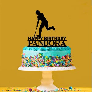 Könnte beinhalten: Schwarze Silhouette eines Hockeyspielers mit Schläger auf einem Kuchen mit blauer Glasur und bunten Streuseln. Der Kuchenaufsatz sagt auch "Happy Birthday Pandora".