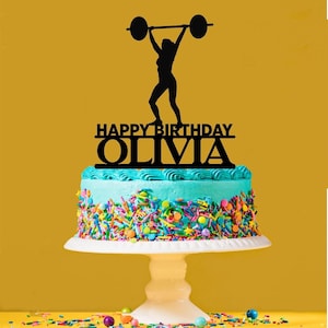 Può includere: Topper per torta in silhouette nera di una persona che solleva un bilanciere con il testo "Happy Birthday Olivia" su una torta glassata blu e bianca.