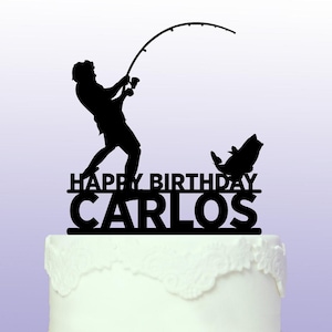 Op de afbeelding: Zwarte silhouet-taarttopper van een persoon die vist met een hengel en een vis. De topper zegt "Happy Birthday Carlos"