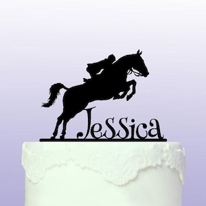 May include: Black silhouette of a horse jumping over a hurdle with a rider on top. The name Jessica is written below the horse. The silhouette is on a white cake.