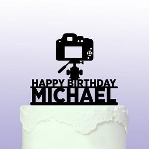 May include: Black and white silhouette of a camera on a tripod with the text "Happy Birthday Michael" below it. The camera is on top of a white cake.