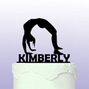 Può includere: Silhouette nera di una persona che fa un ponte con il nome Kimberly scritto sotto. Questo è un topper per torte.