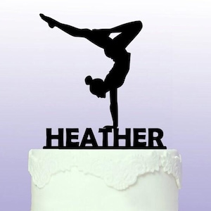 Può includere: Silhouette nera di una ginnasta che fa un handstand su un topper per torta con il nome Heather.