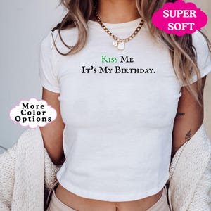 May include: White cropped t-shirt with the text "Kiss Me It's My Birthday." in black lettering. The text "Kiss Me" is in green. The shirt is being modeled by a person.