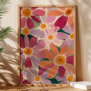 Op de afbeelding: Een ingelijste kunstafdruk met een levendig bloemenpatroon. De kunst toont grote, gestileerde bloemen in roze, oranje en paars met gele centra, tegen een crèmekleurige achtergrond. Het kunstwerk wordt gepresenteerd op een houten kast met een gevlochten mand en een plant.