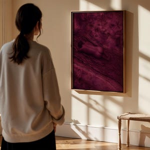 Op de afbeelding: Een rechthoekig wandkunstwerk met een diep magenta en donkerpaarse monochrome sc&egrave;ne van een figuur die op een getextureerd oppervlak ligt. Het kunstwerk is ingelijst in een dunne, lichtgekleurde houten lijst en hangt aan een lichte muur.