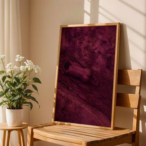 Op de afbeelding: Een rechthoekige ingelijste muurkunstprint met een diep magenta en donkerpaarse illustratie van een figuur die op een getextureerd, golvend oppervlak ligt. Het kunstwerk is gevat in een lichtgekleurde houten lijst met een hoog contrast.