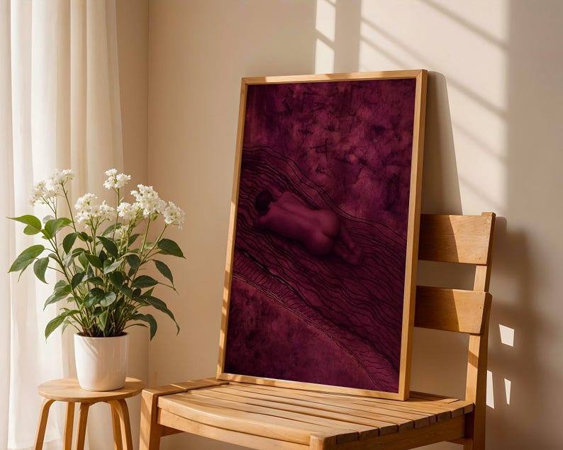Op de afbeelding: Een rechthoekige ingelijste muurkunstprint met een diep magenta en donkerpaarse illustratie van een figuur die op een getextureerd, golvend oppervlak ligt. Het kunstwerk is gevat in een lichtgekleurde houten lijst met een hoog contrast.