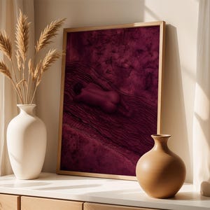Op de afbeelding: Een ingelijste rechthoekige wandkunstprint met een diep magenta achtergrond en een donkere, abstracte figuur die tussen golvende lijnen ligt. De print staat naast een hoge witte keramische vaas met pampasgras en een kleine beige vaas.