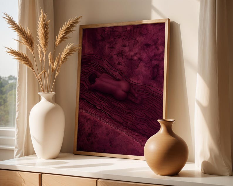 Op de afbeelding: Een ingelijste rechthoekige wandkunstprint met een diep magenta achtergrond en een donkere, abstracte figuur die tussen golvende lijnen ligt. De print staat naast een hoge witte keramische vaas met pampasgras en een kleine beige vaas.