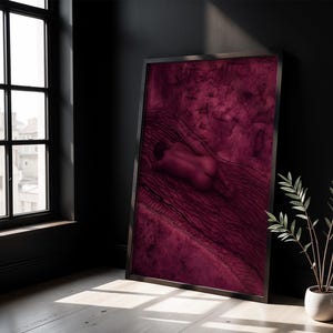 Op de afbeelding: Een ingelijste verticale wandkunstprint met een diep magenta en donkerrode monochrome illustratie van een menselijk figuur dat op een getextureerd oppervlak ligt met golvende lijnpatronen en verspreide donkere markeringen.