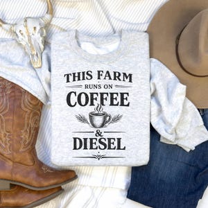 May include: A light gray sweatshirt with the text "THIS FARM RUNS ON COFFEE & DIESEL" in black. The design includes a coffee cup graphic. The sweatshirt is displayed with a cowboy hat, boots, and a cow skull.
