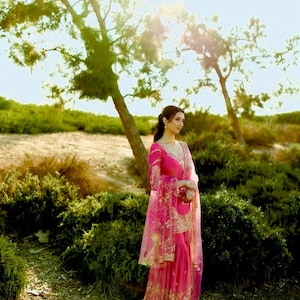 Puede incluir: Una mujer con un vestido bordado en rosa y dorado con un dupatta transparente. Está de pie en un campo con árboles y arbustos verdes en el fondo.