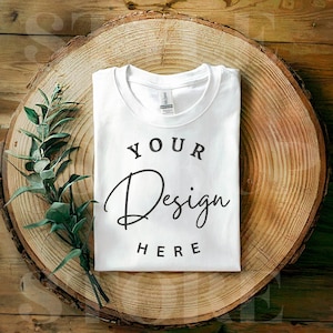 Puede incluir: Maqueta de camiseta blanca sobre un fondo de madera con vegetación. La camiseta tiene el texto "YOUR DESIGN HERE" impreso.