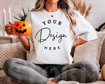 May include: White t-shirt with the text "YOUR Design HERE" in black. An orange pumpkin-shaped mug with a carved jack-o'-lantern face is held in the hand. The person is wearing gray jeans and sitting on a couch.