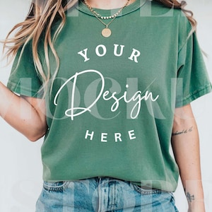 Puede incluir: Una camiseta verde con las palabras "YOUR Design HERE" en escritura blanca. La camiseta se combina con pantalones cortos de mezclilla y collares dorados. La camiseta tiene mangas cortas y cuello redondo.