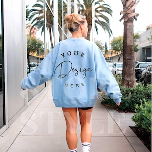Puede incluir: Una sudadera azul claro con el texto "YOUR Design HERE" impreso en la espalda. La sudadera la lleva puesta una persona que camina por una acera.