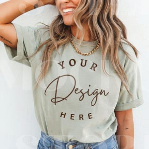 May include: A light green t-shirt with the text "YOUR Design HERE" in a brown script font. The shirt is paired with blue denim jeans and a gold chain necklace. The model is smiling.