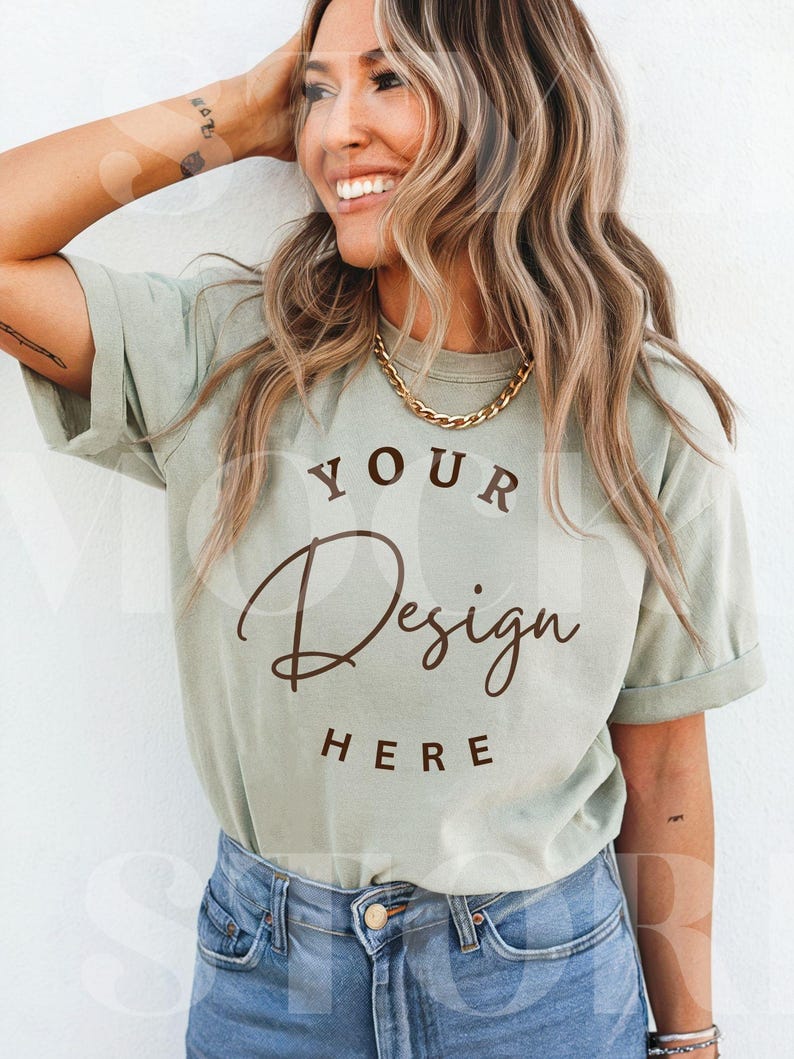 May include: A light green t-shirt with the text "YOUR Design HERE" in a brown script font. The shirt is paired with blue denim jeans and a gold chain necklace. The model is smiling.