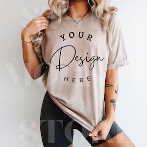 Peut inclure: Un t-shirt beige avec le texte "YOUR Design HERE" imprimé sur le devant. Le t-shirt est porté par une personne avec un tatouage sur le bras.