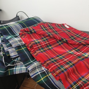 以下が含まれることがあります： 赤、緑、青、黄色のタータンチェック柄の折り畳まれた生地。生地は積み重ねられており、クラシックなタータンパターンを示しています。赤いタータンチェック柄が最も目立ち、鮮やかな色と複雑なデザインが特徴です。