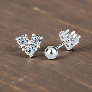 May include: Two silver stud earrings. One earring features three clear, round-cut cubic zirconia stones arranged in a triangular shape. The other earring has a small silver ball and a decorative backing.