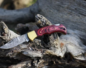 Puede incluir: Un cuchillo plegable de bolsillo con una hoja de acero estampada y un mango de madera rojo y marrón.