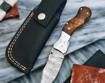 Puede incluir: Una navaja plegable con una hoja estampada y un mango de madera con remaches de latón. El cuchillo se combina con una funda de cuero negro con costuras doradas y un cierre de botón plateado. La hoja tiene un patrón arremolinado único.