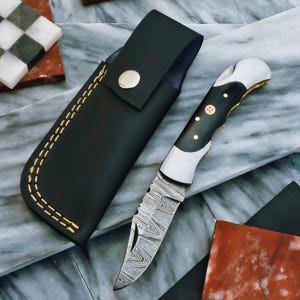 May include: A black leather knife sheath with a gold stitch detail and a black and silver pocket knife with a damascus steel blade and a black handle with a red cross design.