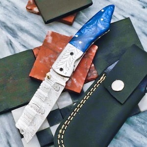 以下が含まれることがあります： 模様のある刃と青と白のハンドルが付いた折りたたみナイフ。ハンドルには白いドットのアクセントが付いています。ナイフは、スナップ留めが付いた黒い革の鞘の隣にあります。ブレードにはユニークな波状のパターンがあります。