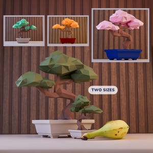 May include: Two origami bonsai trees in different sizes, one green and one brown, with a yellow banana on a white surface. The text "TWO SIZES" is visible in the image.