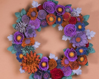 May include: A handcrafted felt flower wreath featuring a variety of colorful blooms in shades of purple, orange, red, and blue. The wreath is adorned with green and gray felt leaves, creating a vibrant and textured floral arrangement for home decor.