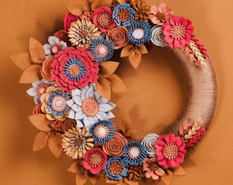May include: A handcrafted floral wreath featuring a variety of felt flowers in shades of pink, blue, and beige, accented with brown leaves. The wreath's base is wrapped in a natural-colored twine, creating a rustic and decorative piece.