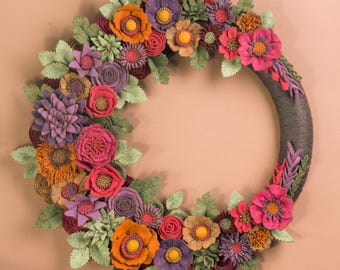 May include: A handmade floral wreath featuring a variety of felt flowers in shades of orange, purple, pink, and red, with green leaves. The wreath is circular and has a dark gray base, perfect for home decor.