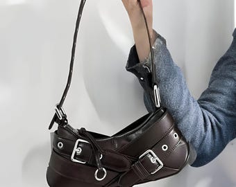 May include: A brown leather handbag with silver buckles and a strap. The bag has a unique shape and is designed with a modern aesthetic.