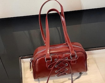 May include: A red leather satchel handbag with a lace-up detail on the front. The bag has two top handles and a detachable shoulder strap.