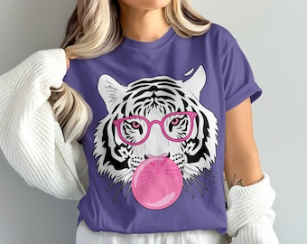 May include: Purple t-shirt featuring a white tiger face with black stripes, wearing pink glasses and blowing a pink bubble. The graphic tee is a fun and stylish design, perfect for casual wear.