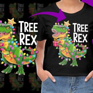 May include: Black t-shirt featuring a green dinosaur wrapped in Christmas lights, holding a gift. The text "TREE REX" is printed in white above the dinosaur. A gold star is on the dinosaur's head.