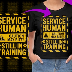 Puede incluir: Camiseta negra con un gráfico amarillo y negro que dice "SERVICE HUMAN CAUTION MAY BITE! STILL IN TRAINING." El diseño incluye huellas de patas y un símbolo de advertencia.